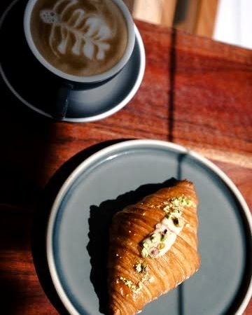 Croissant de pistacho abierto junto a un cappuccino sobre una mesa de madera oscura.
