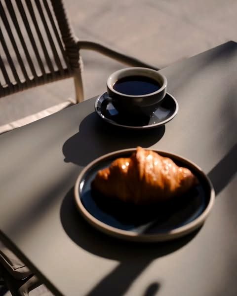 Un café solo y un croissant sobre una mesa al sol, junto a una silla de la terraza.