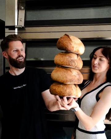 Dos panaderos de Esquina Bakery sostienen una torre de panes redondos delante del horno.